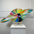 A Damien Hirst colorful abstract ringed planet sculpture made of painted wood, displayed on a small wooden easel stand atop a white pedestal against a light gray paneled wall.