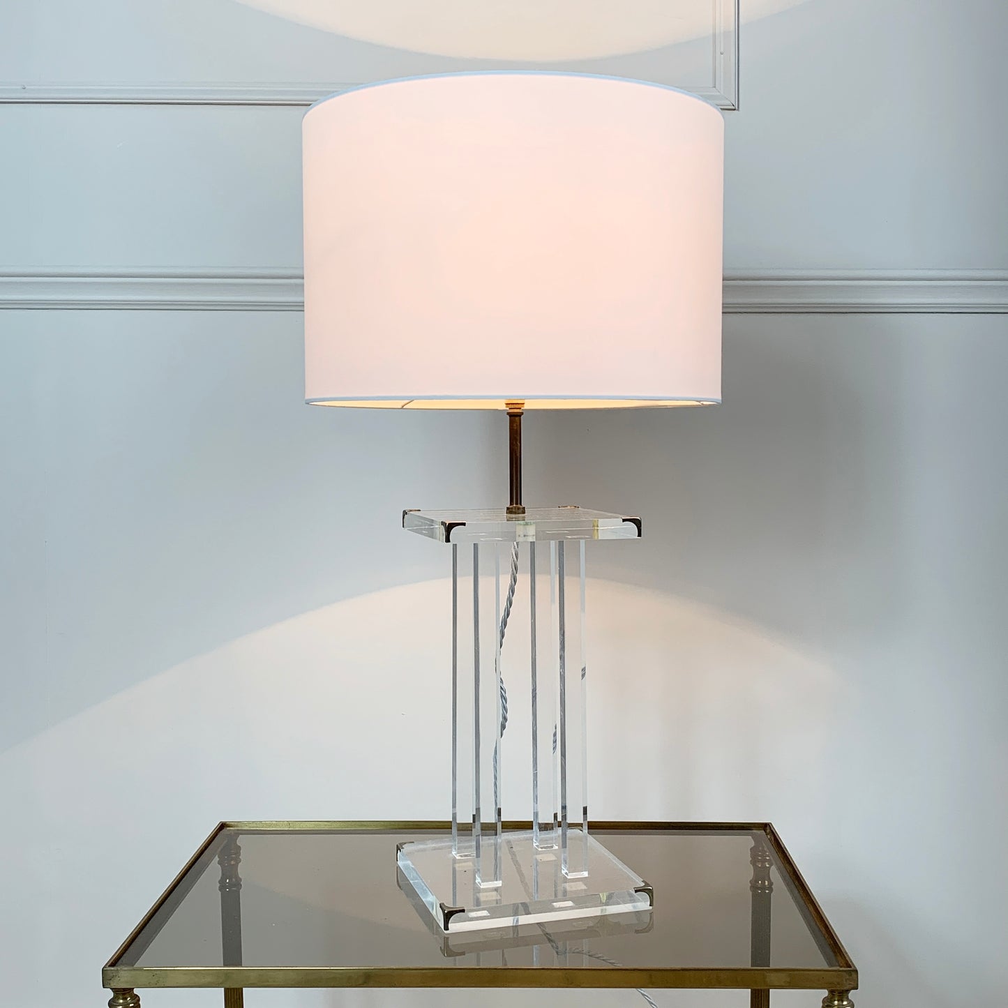 A table lamp with a large white drum shade, a clear acrylic geometric base, and a brass-toned stem, shown lit on a mirrored side table against a light gray wall.