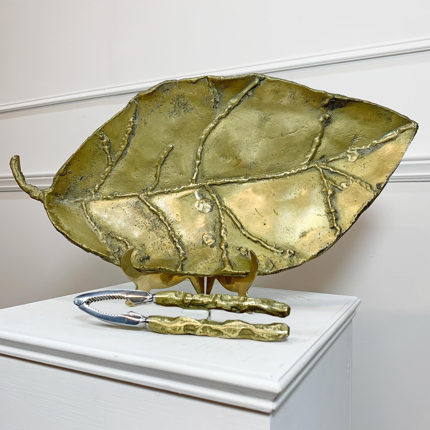 A large David Marshall  leaf-shaped serving tray or dish in a metallic gold finish with raised vein details, displayed on a white pedestal with a pair of matching gold-handled nutcrackers in front.
