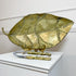 A large David Marshall  leaf-shaped serving tray or dish in a metallic gold finish with raised vein details, displayed on a white pedestal with a pair of matching gold-handled nutcrackers in front.
