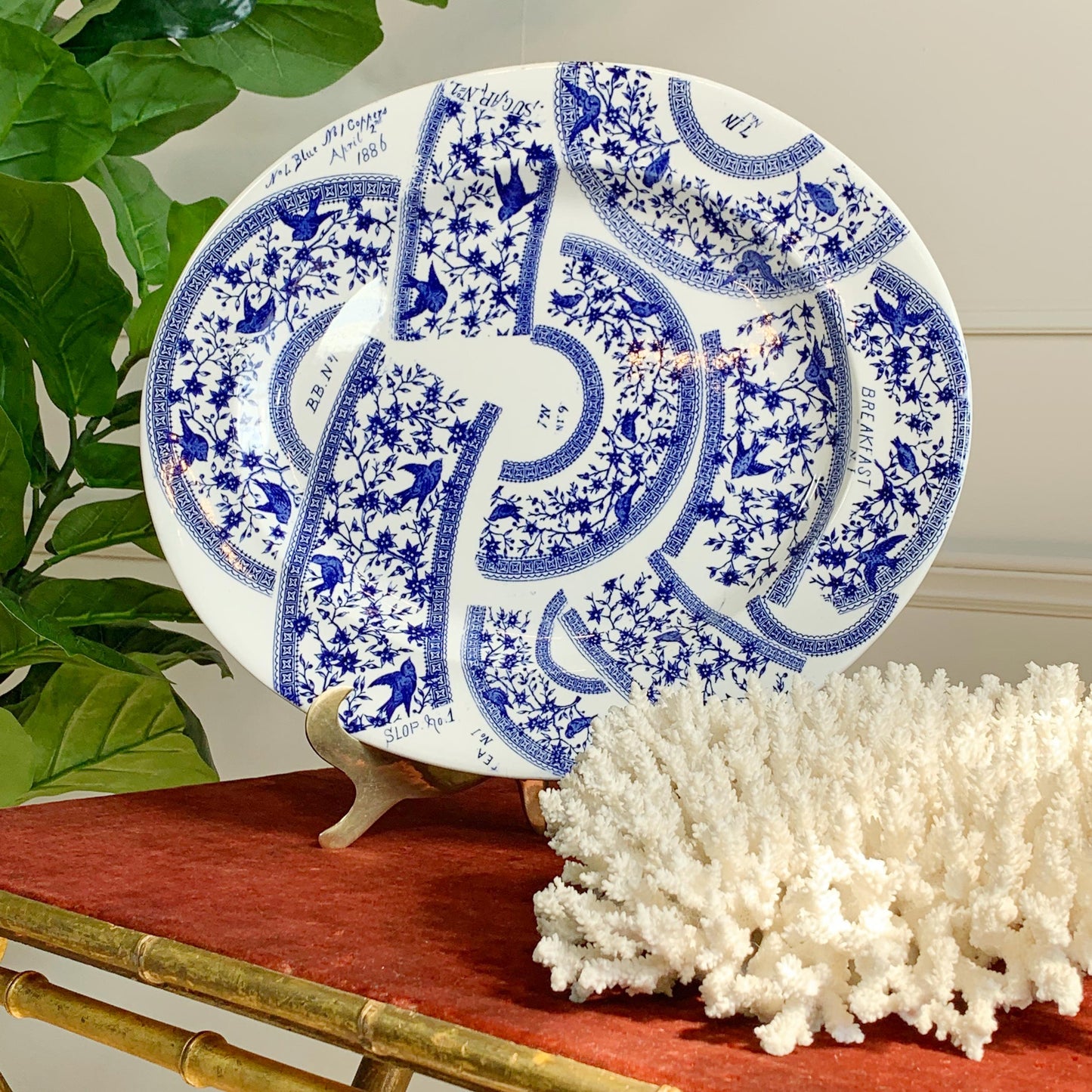 An oval white ceramic plate with an intricate blue floral and bird pattern, displayed on a small stand on a red tablecloth next to a white coral piece and green leaves.