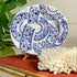 An oval white ceramic plate with an intricate blue floral and bird pattern, displayed on a small stand on a red tablecloth next to a white coral piece and green leaves.