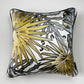 A square decorative silk pillow featuring a stylized tropical palm leaf pattern in gold, black, and gray on a white background, with a black piped edge.
