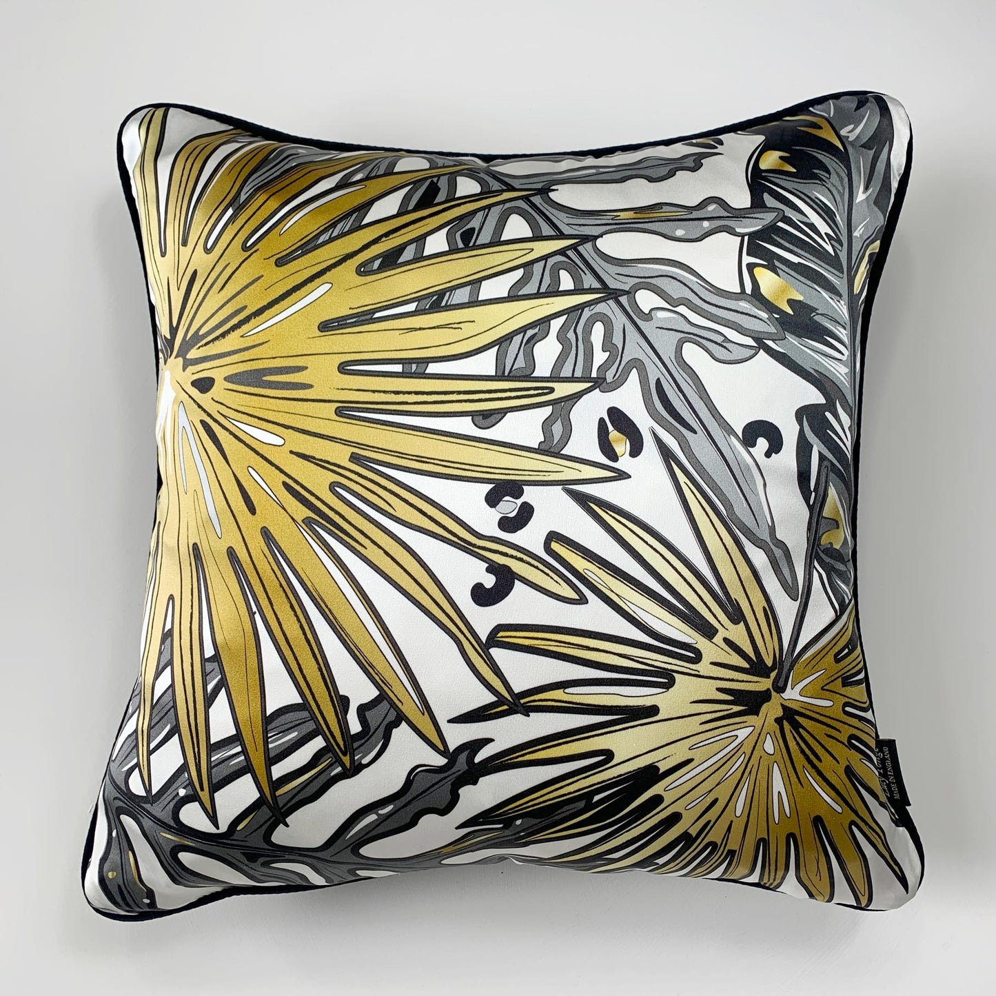 A square decorative silk pillow featuring a stylized tropical palm leaf pattern in gold, black, and gray on a white background, with a black piped edge.