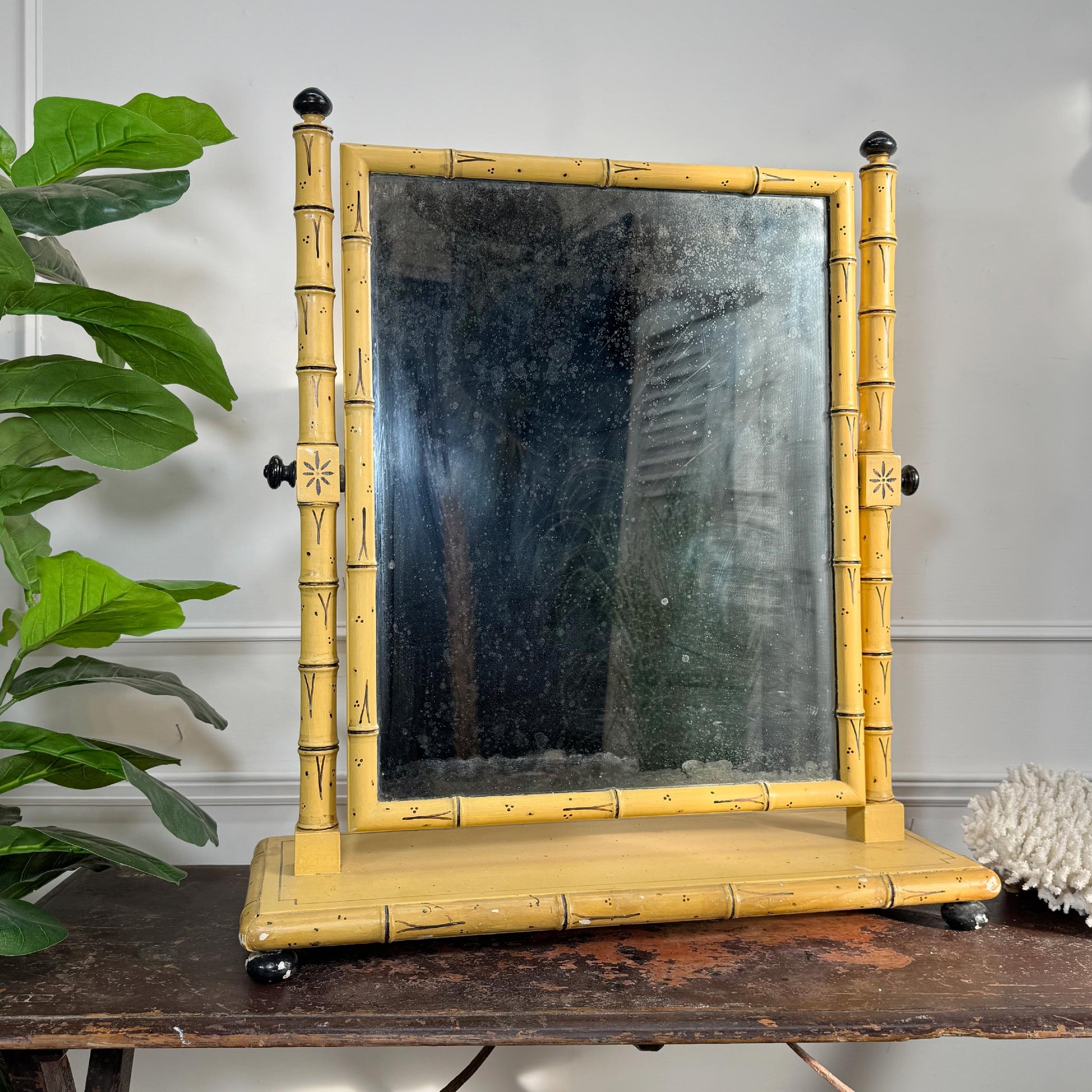 A regency style painted faux bamboo frame mirror with a yellow and black painted finish, resting on a rustic wooden table next to a green plant.