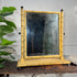 A regency style painted faux bamboo frame mirror with a yellow and black painted finish, resting on a rustic wooden table next to a green plant.