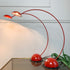A mid-century modern table lamp with two adjustable dome-shaped shades on curved red metal arms, mounted on a matching red base, shown on a glass tabletop against a white paneled wall.