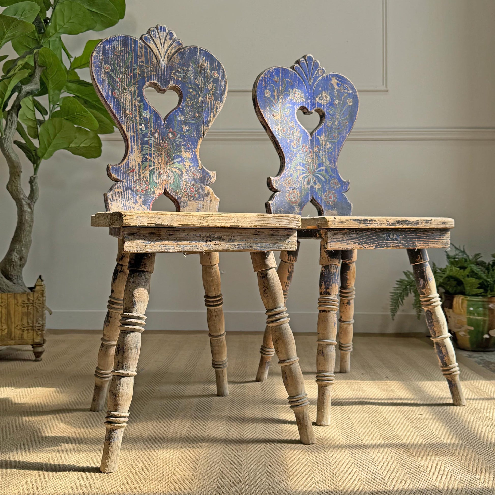 Pair of blue hand painted rustic wooden folk chairs with decorative backs in a room with plants.