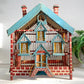 A decorative miniature house-shaped birdcage with a blue painted roof, red-and-white brick patterned sides, and yellow perches inside, photographed against a plain light background with a plant partially visible on the right.