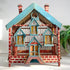 A decorative miniature house-shaped birdcage with a blue painted roof, red-and-white brick patterned sides, and yellow perches inside, photographed against a plain light background with a plant partially visible on the right.