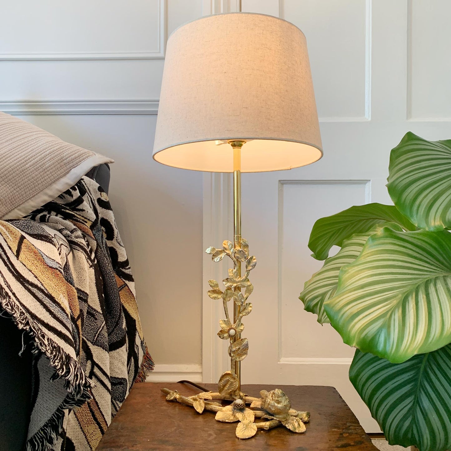 A Fondica Gold Bird & Flowers Table Light, by Regis Dho with a warm beige fabric drum shade and an ornate gold-toned floral base, styled on a wooden table next to a green plant and a patterned throw blanket.
