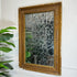 An antique rectangular wall mirror with a distressed, ornate gold frame and foxed crackled mirror plate, mounted on a white wall with a green plant visible on the left.
