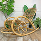 A natural rattan and bamboo rocking chair, styled on a rustic wooden floor with potted green plants against a white paneled wall.