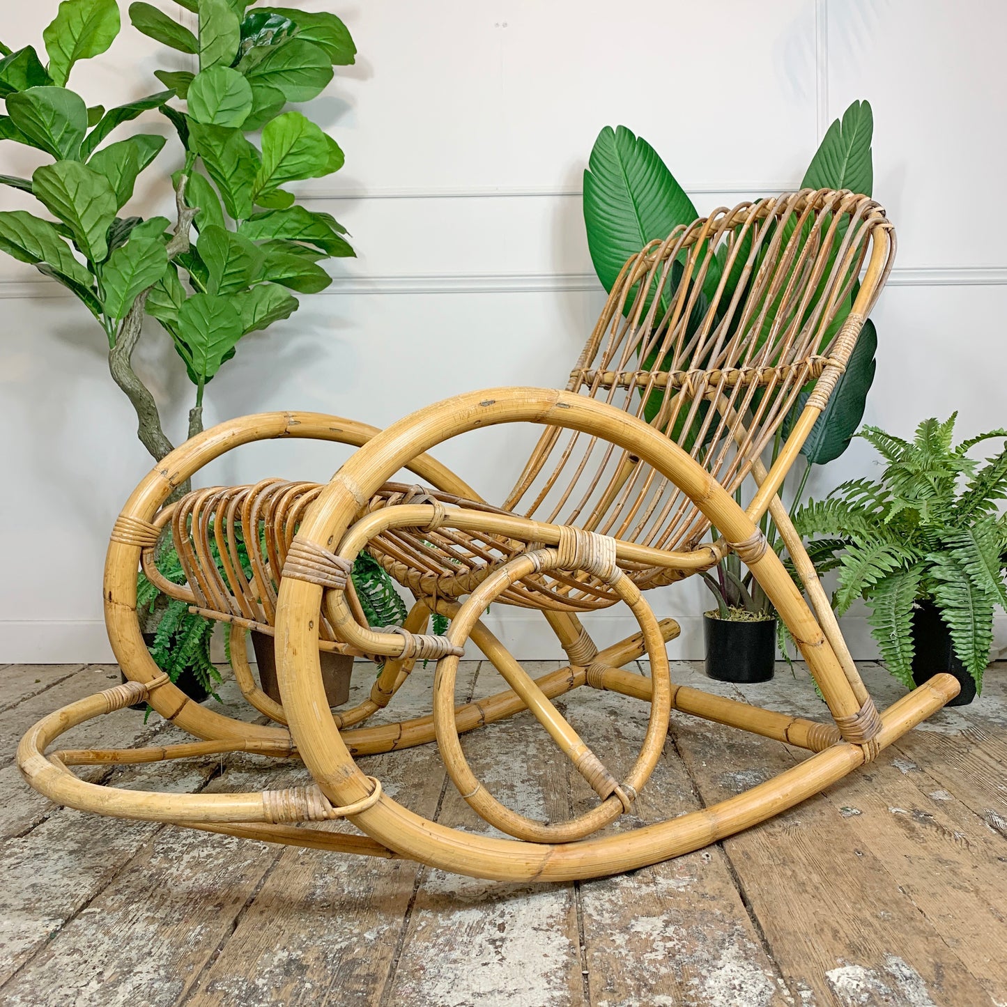 A natural rattan and bamboo rocking chair, styled on a rustic wooden floor with potted green plants against a white paneled wall.