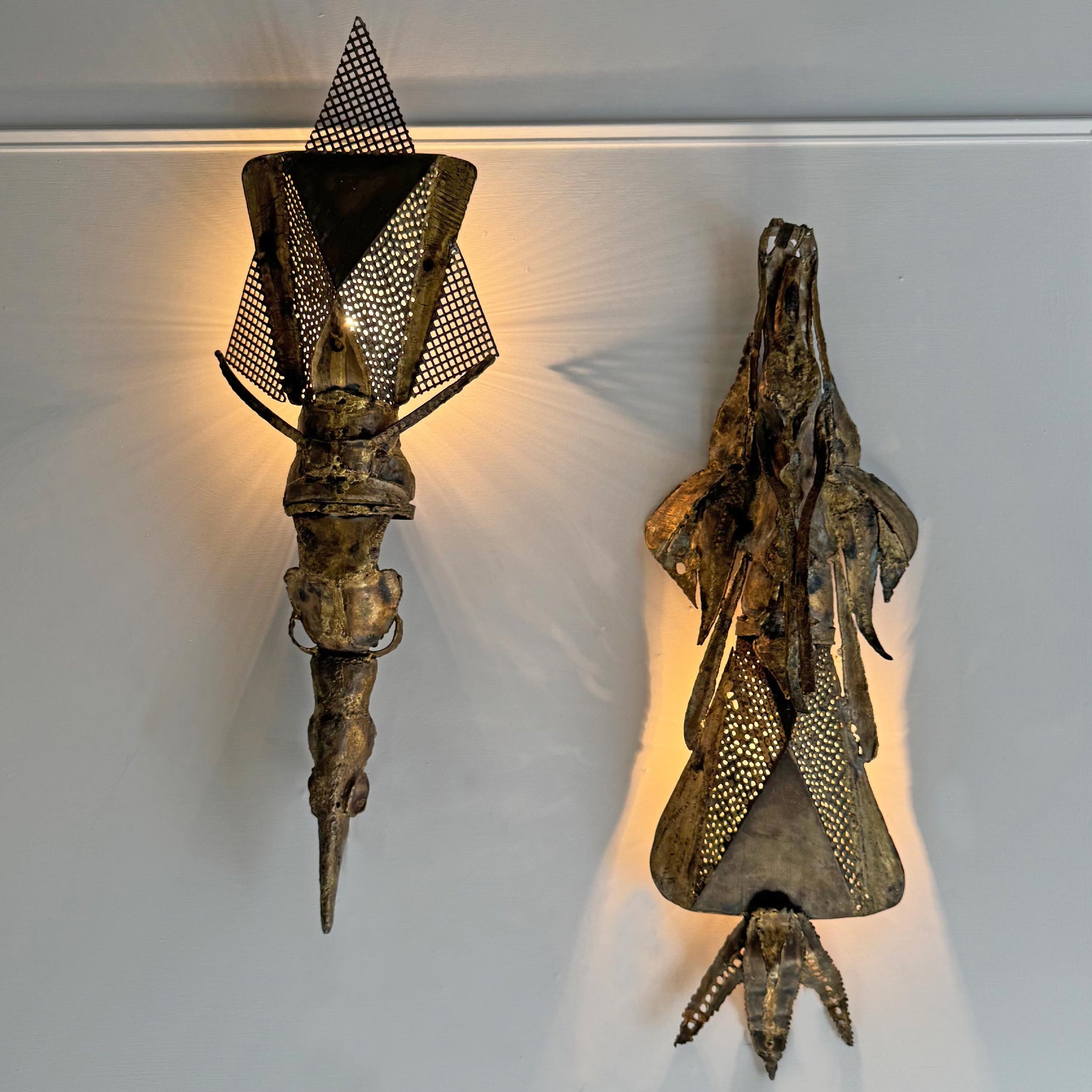 Two sculptural wall sconces Francois Melin with an organic, metallic form in a distressed bronze finish, mounted on a white wall with warm light radiating from behind them.