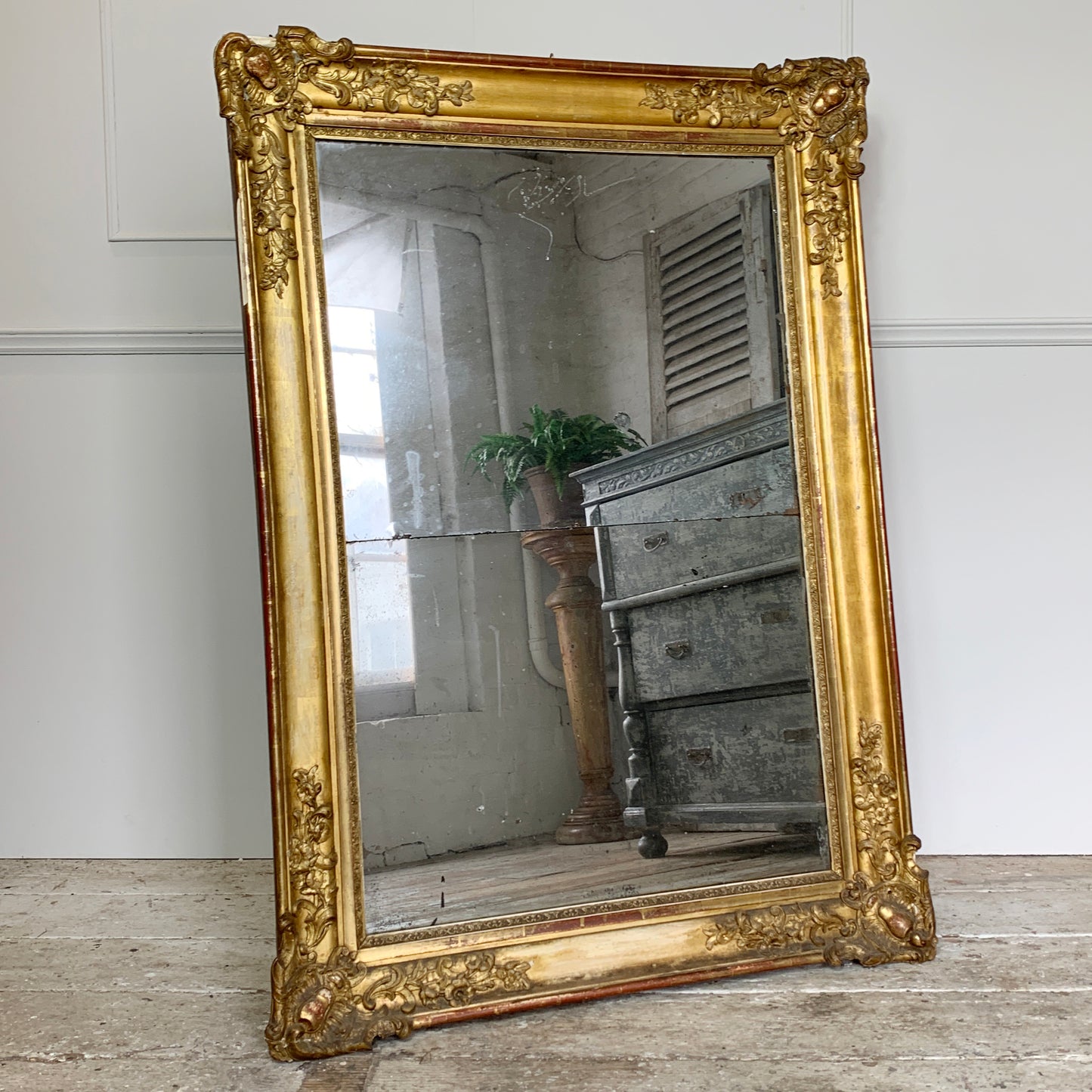 An ornate antique french split plate wall mirror with a wide carved gold frame, leaning against a white wall on a rustic wooden floor. The mirror reflects a room interior with a gray wooden chest and a potted plant.