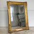 An ornate antique french split plate wall mirror with a wide carved gold frame, leaning against a white wall on a rustic wooden floor. The mirror reflects a room interior with a gray wooden chest and a potted plant.