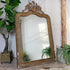 An ornate 19th Century French Crested Gold Gesso Mirror with a distressed gold and brown carved frame, leaning against a white paneled wall on a rustic wooden surface, flanked by potted plants.