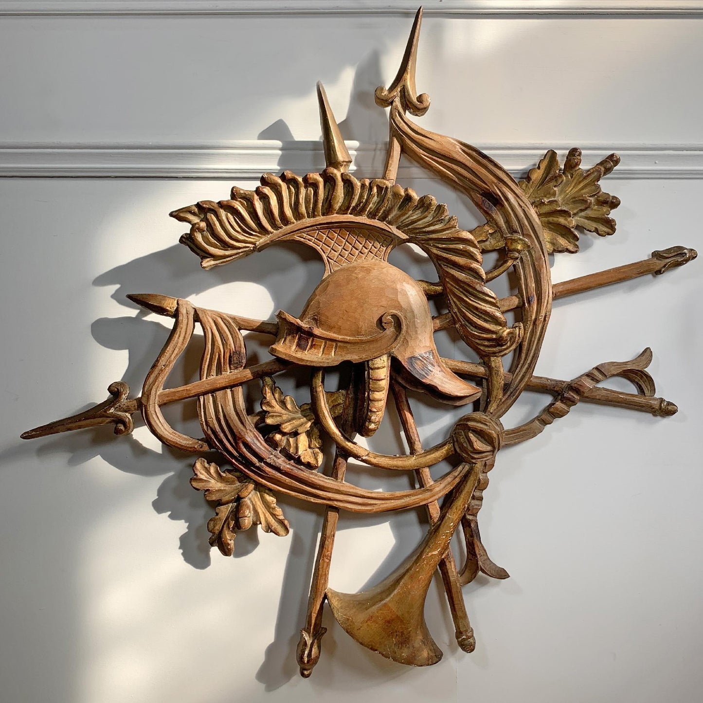 An antique carved wooden wall sculpture featuring a helmet, trident, leaf motifs, and crossed spear-like elements, displayed against a white wall with natural sunlight casting shadows.
