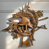 An antique carved wooden wall sculpture featuring a helmet, trident, leaf motifs, and crossed spear-like elements, displayed against a white wall with natural sunlight casting shadows.