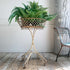 A green fern plant displayed in a round metal planter on a distressed cream-colored metal stand, set on a rustic wooden floor against a white paneled wall.