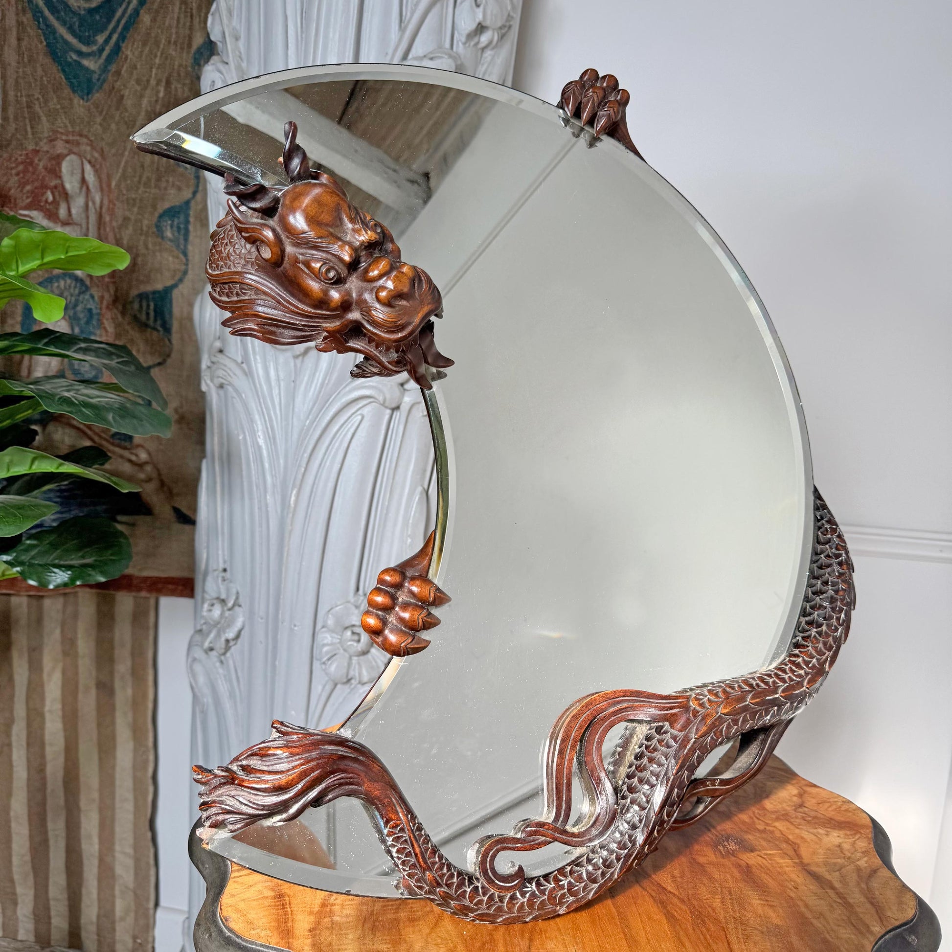 A Gabrielle Viardot ornate round wall mirror with a carved bronze dragon head at the top and a dragon body wrapping around the base, displayed on a wooden surface in an indoor setting.