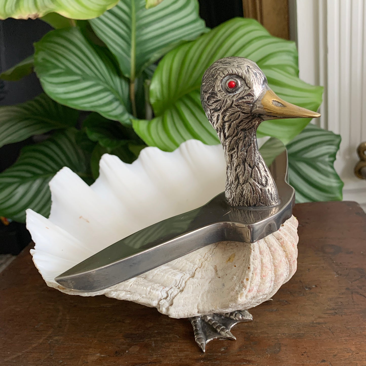 A decorative gabriella binazzi duck-shaped osculpture with a silver head and beak, a red eye, and a gold beak tip, mounted on a white scalloped clam shell over a wooden surface, with green leafy plants in the background.