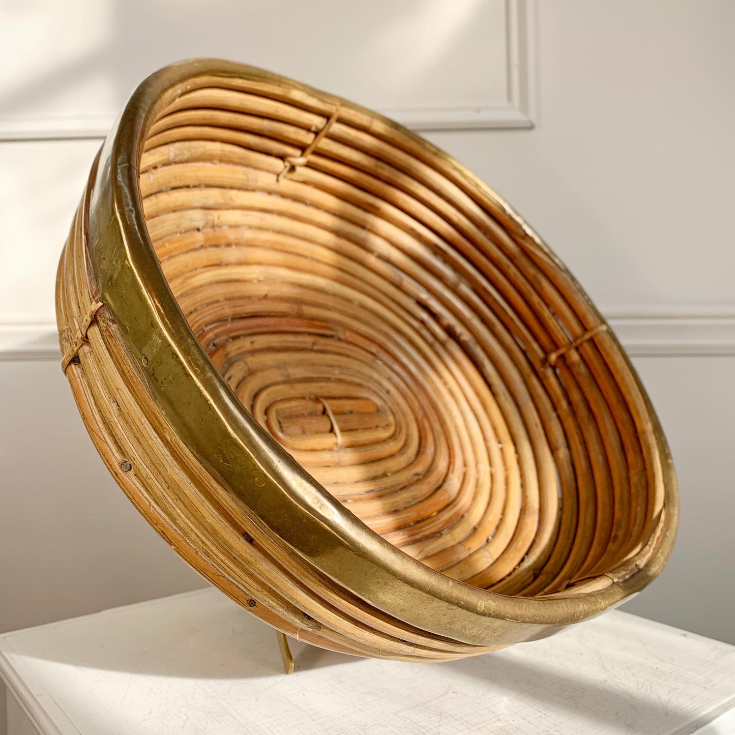 A round woven rattan Crespi basket with a natural tan color and a polished brass rim, displayed on a white surface against a white paneled wall.