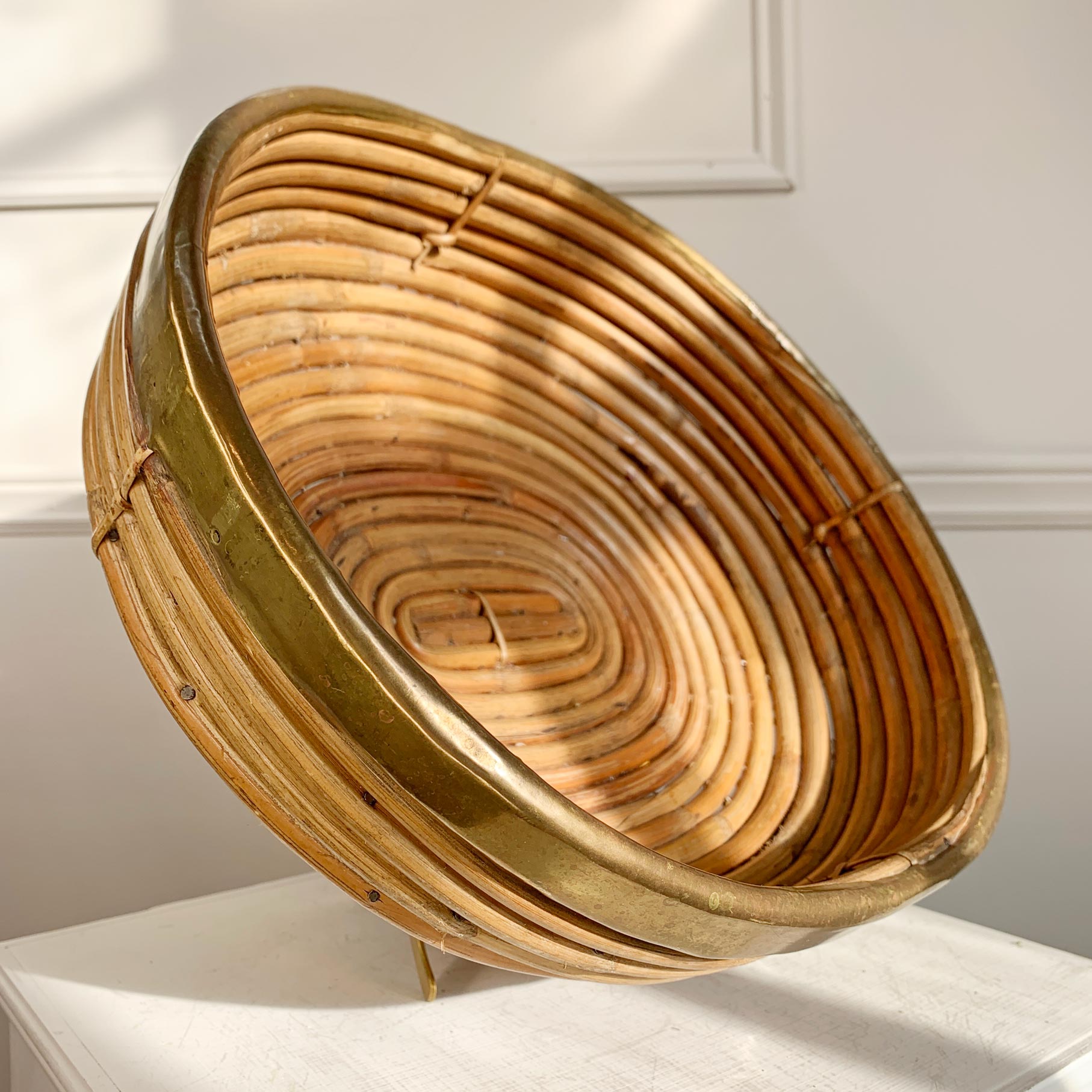 A round woven rattan Crespi basket with a natural tan color and a polished brass rim, displayed on a white surface against a white paneled wall.
