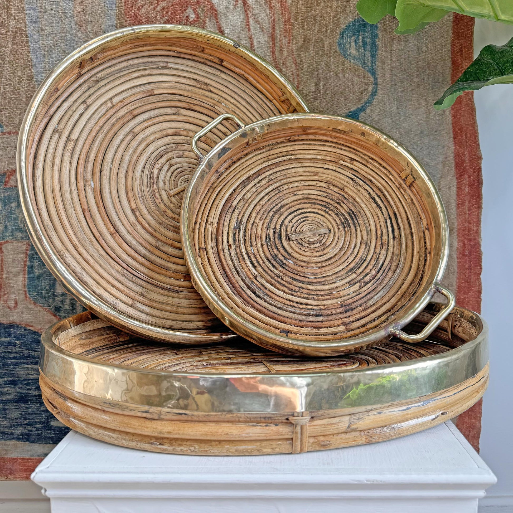 Three round Gabriella Crespi style woven rattan trays with brass-colored metal rims and handles, stacked on a white pedestal against a rustic patterned backdrop.