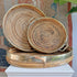 Three round Gabriella Crespi style woven rattan trays with brass-colored metal rims and handles, stacked on a white pedestal against a rustic patterned backdrop.
