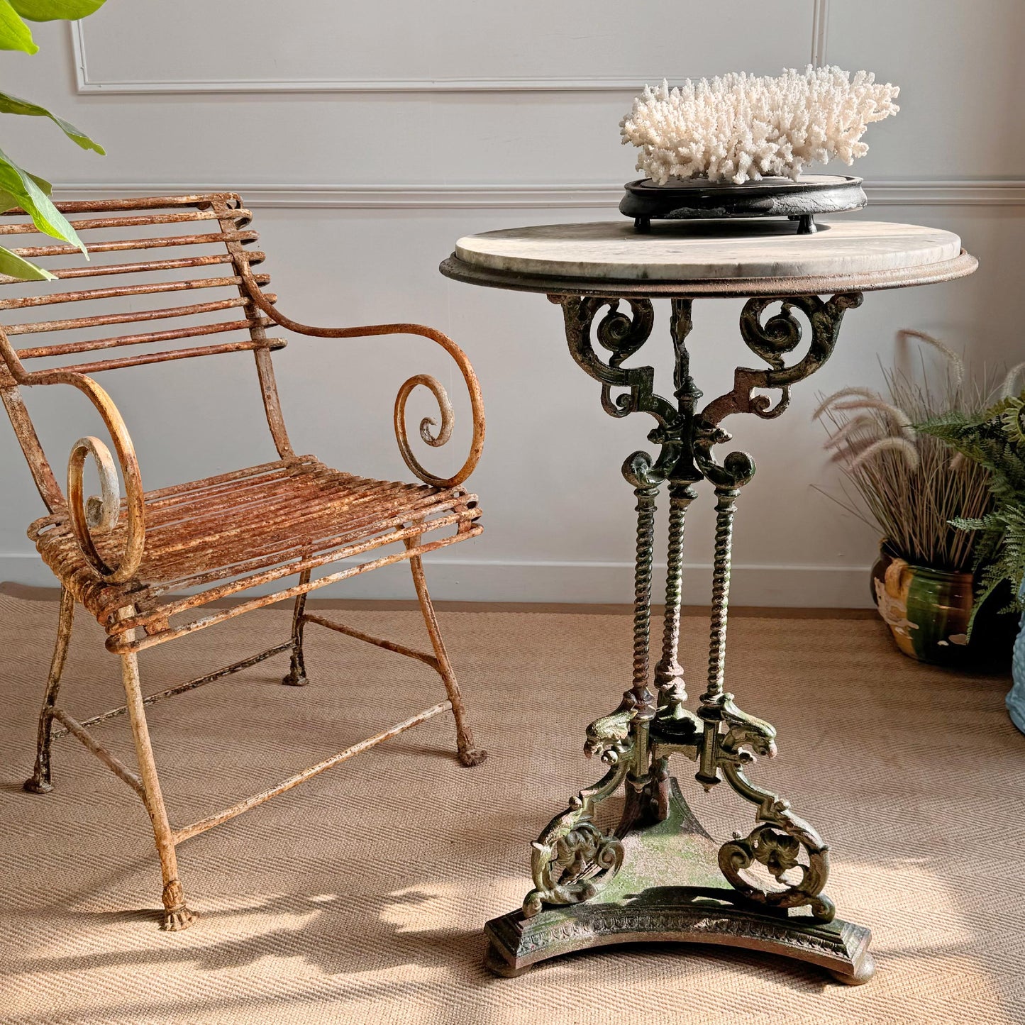An ornate round antique English cast iron metal pedestal garden table with a marble top, stands beside a n antique Arras metal armchair in a styled interior setting with a marble-look top and decorative coral on top.