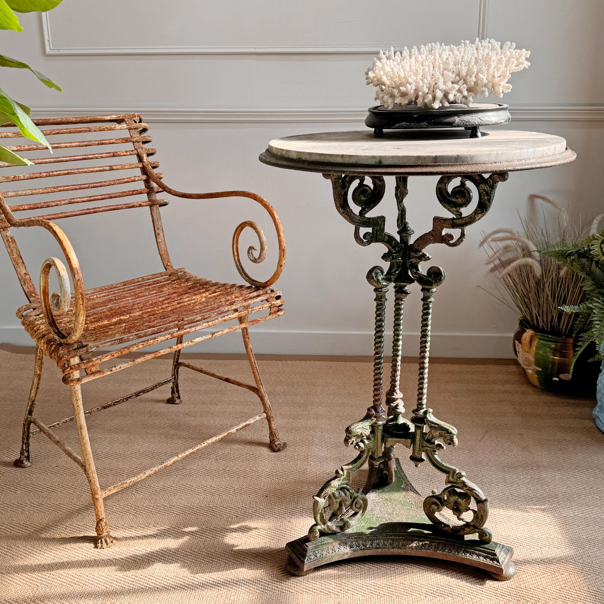 An ornate round antique English cast iron metal pedestal garden table with a marble top, stands beside a n antique Arras metal armchair in a styled interior setting with a marble-look top and decorative coral on top.