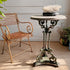 An ornate round antique English cast iron metal pedestal garden table with a marble top, stands beside a n antique Arras metal armchair in a styled interior setting with a marble-look top and decorative coral on top.