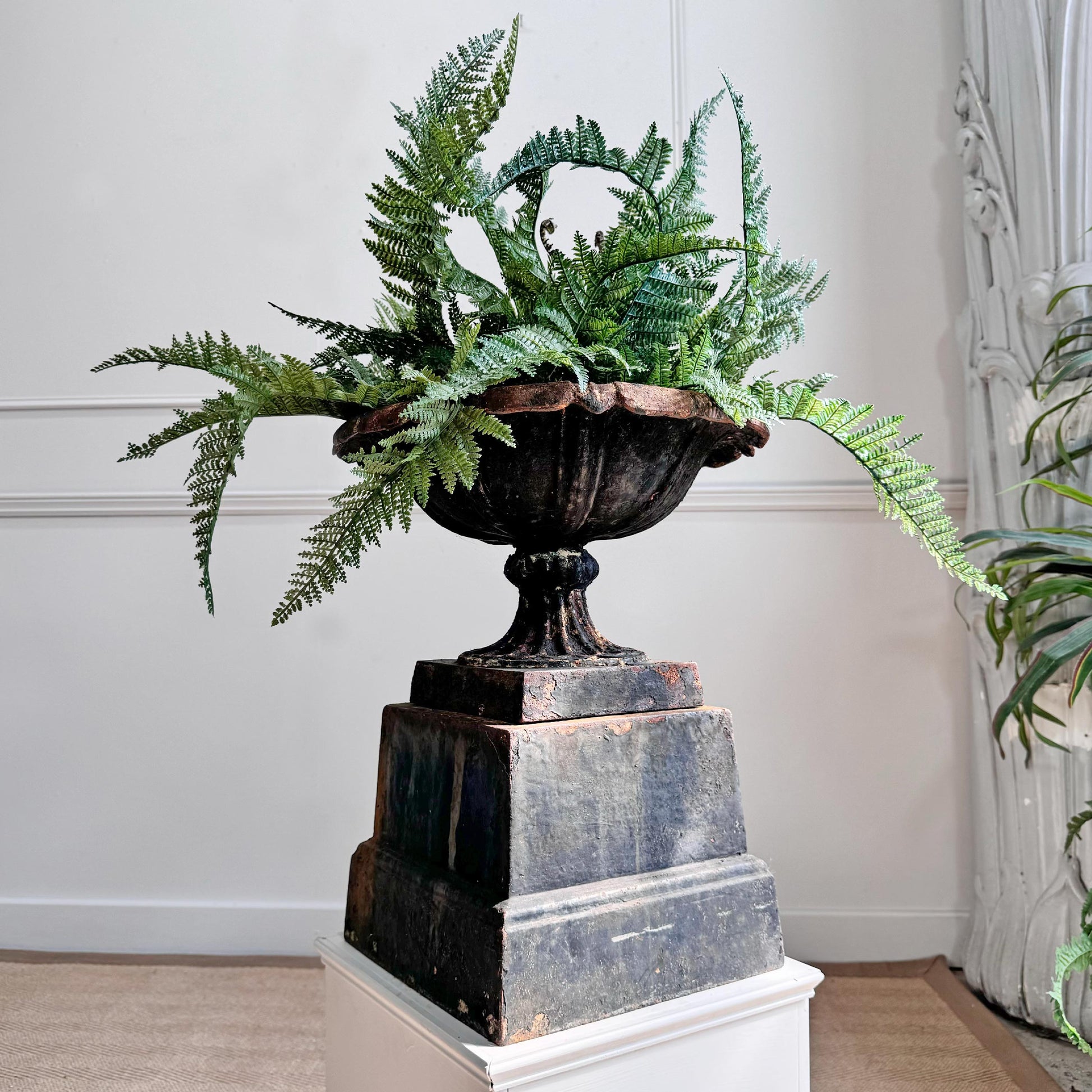 An ornate dark metal urn planter filled with lush green ferns, displayed on a tiered dark pedestal atop a white plinth in an indoor setting.