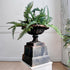 An ornate dark metal urn planter filled with lush green ferns, displayed on a tiered dark pedestal atop a white plinth in an indoor setting.
