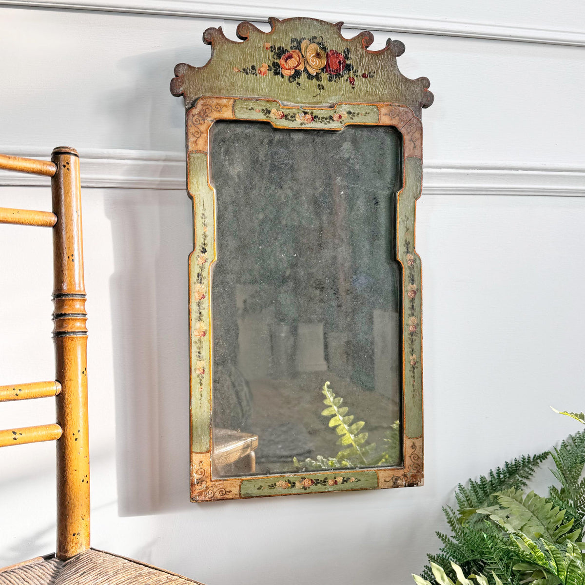 An antique Georgian rectangular wall mirror with a distressed green and gold painted frame, featuring floral details and a decorative crest at the top, shown hanging on a white wall next to a wooden chair and green plants.