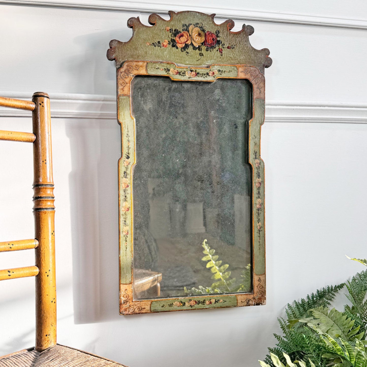 An antique Georgian rectangular wall mirror with a distressed green and gold painted frame, featuring floral details and a decorative crest at the top, shown hanging on a white wall next to a wooden chair and green plants.