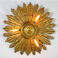 A large, ornate antique wooden gold sunburst ceiling light fixture with multiple textured rays and three visible glowing bulbs, mounted against a plain white wall.