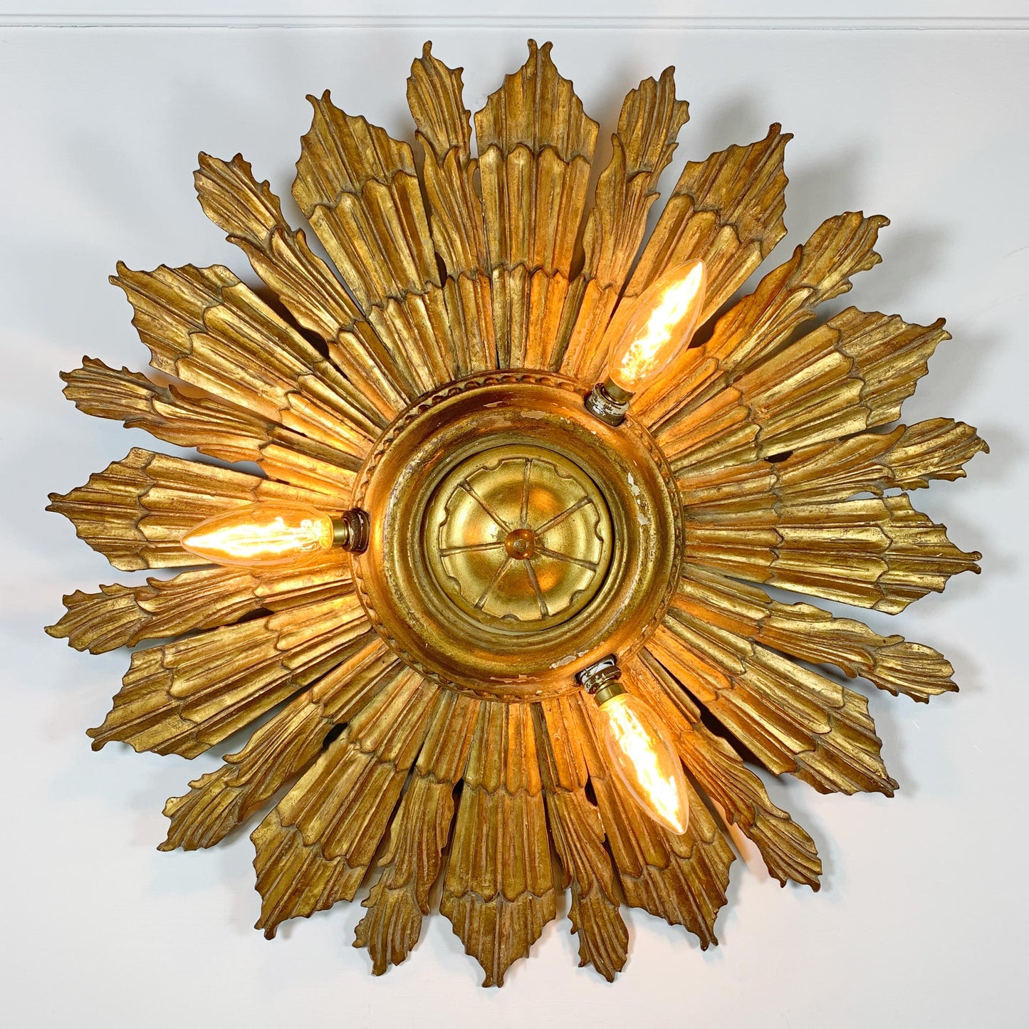 A large, ornate antique wooden gold sunburst ceiling light fixture with multiple textured rays and three visible glowing bulbs, mounted against a plain white wall.