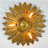 A large, ornate antique wooden gold sunburst ceiling light fixture with multiple textured rays and three visible glowing bulbs, mounted against a plain white wall.