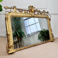 An ornate antique gold mirror with a highly decorative carved frame, leaning against a white paneled wall on a beige carpet. The mirror reflects potted ferns and a window, and a small green plant is visible in the upper left corner.