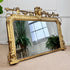 An ornate antique gold mirror with a highly decorative carved frame, leaning against a white paneled wall on a beige carpet. The mirror reflects potted ferns and a window, and a small green plant is visible in the upper left corner.