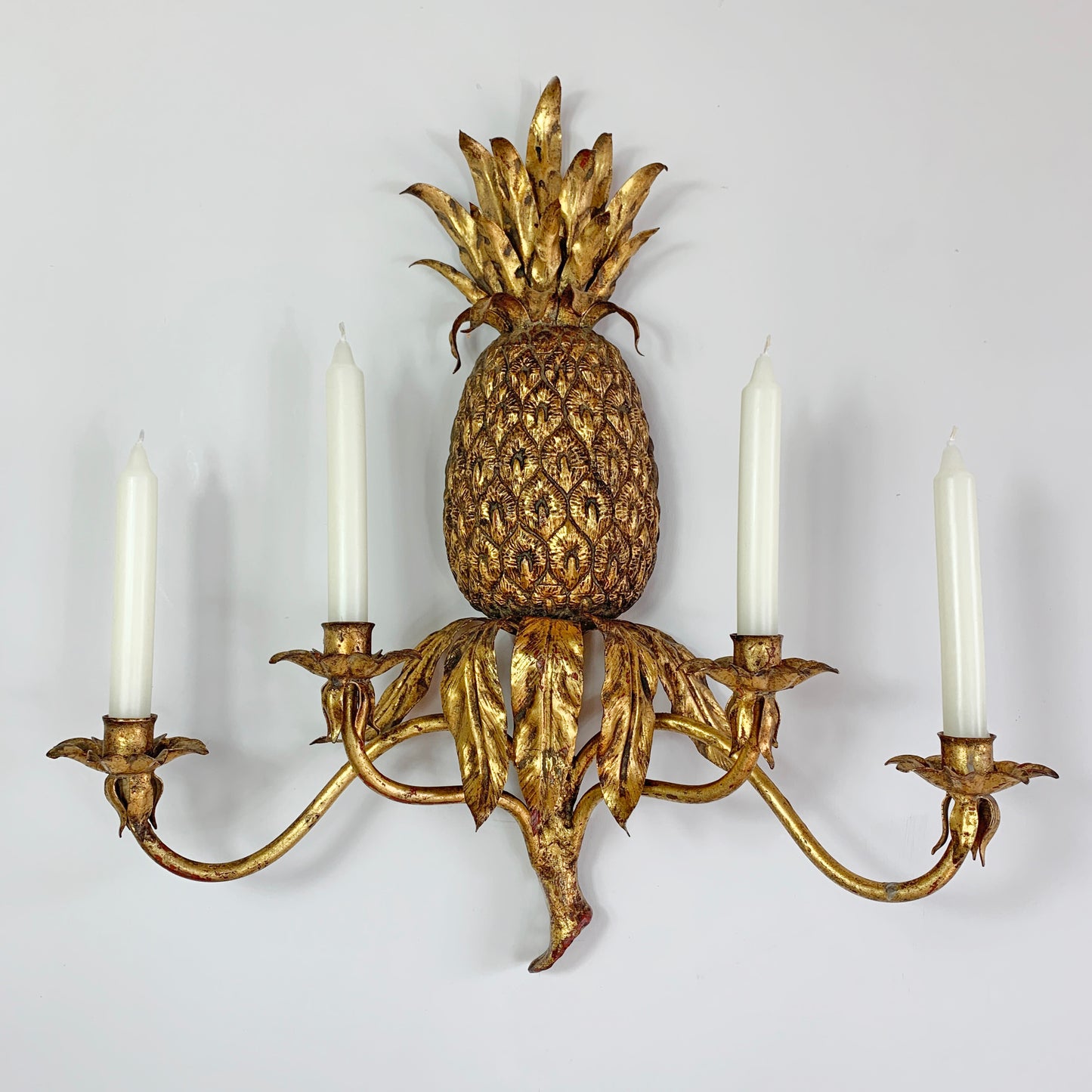 A decorative wall sconce shaped like a pineapple, finished in an antique gold tone, with four white candle-style lights extending from leafy arms.