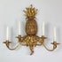 A decorative wall sconce shaped like a pineapple, finished in an antique gold tone, with four white candle-style lights extending from leafy arms.