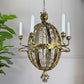 An ornate brass candle chandelier with a spherical cage design, five candle-style arms, and white faux candles, shown hanging against a light wall with a green plant on the left.