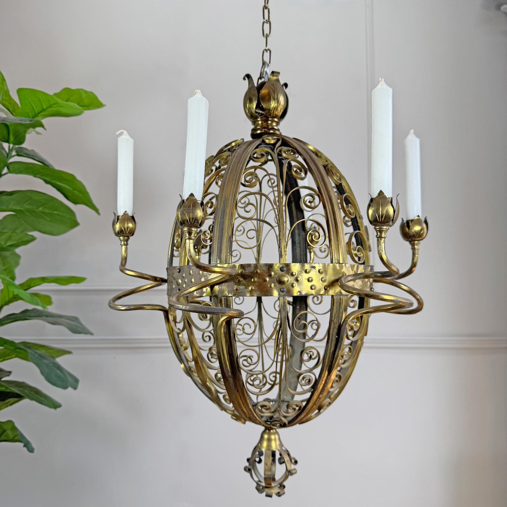 An ornate brass candle chandelier with a spherical cage design, five candle-style arms, and white faux candles, shown hanging against a light wall with a green plant on the left.