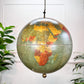 Vintage hanging globe lamp with a detailed map design showing Africa and surrounding continents in muted green, yellow, and orange tones, suspended by a chain against a white paneled wall with plants at the sides.