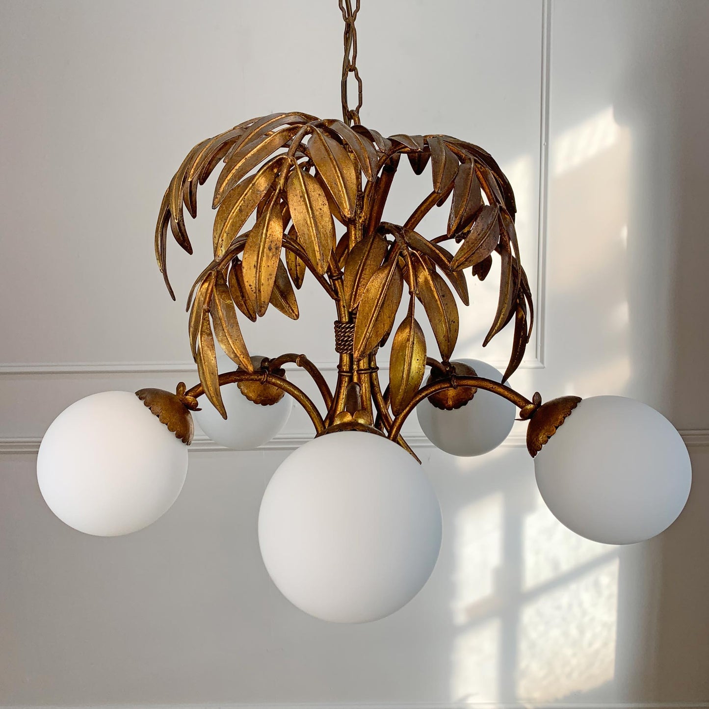 A vintage Hans Kogl gilt palm tree chandelier with a distressed bronze-gold metal frame and five white spherical glass shades, shown hanging against a white paneled wall with soft window light.