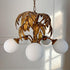A vintage Hans Kogl gilt palm tree chandelier with a distressed bronze-gold metal frame and five white spherical glass shades, shown hanging against a white paneled wall with soft window light.